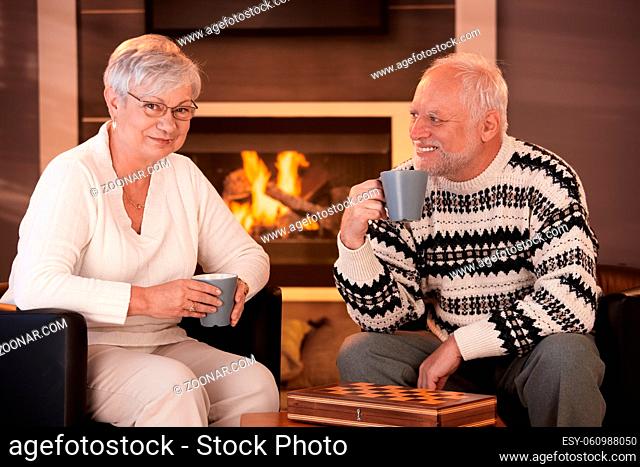 Happy senior couple on winter night at home, sitting in front of fireplace in cosy living room, having tea