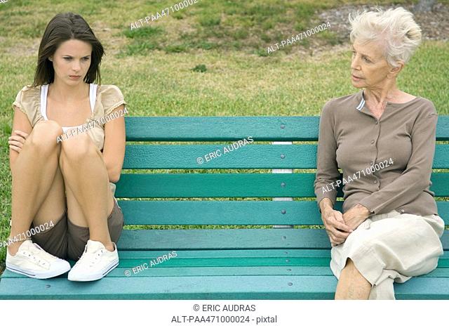 Teenage girl sitting apart from grandmother on bench, arms folded, looking down