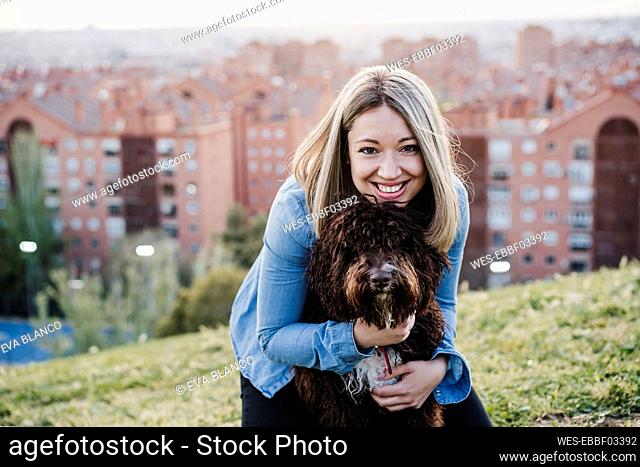 Smiling woman with blond hair embracing Spanish Water Dog on grass during sunset