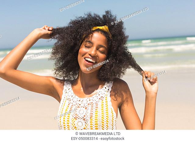 Front view of happy young African American woman with eyes closed playing with hair standing on the beach. She is smiling