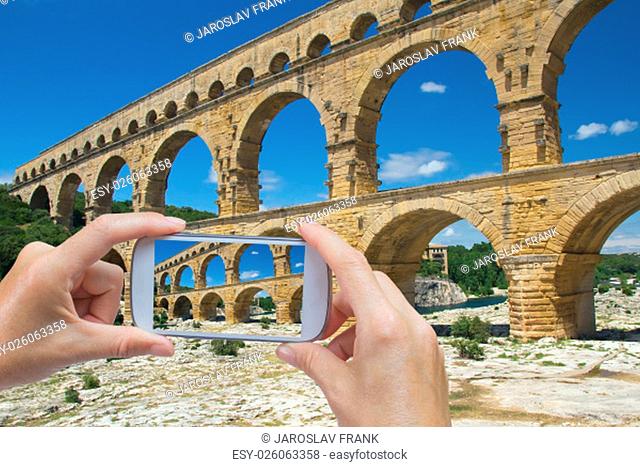 In the bottom left of the photo are hands holding smart phone, whose screen contains photo of Pont du Gard. Background of the photo contains also Pont du Gard