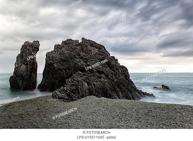 Beach at Monterosso al Mare: imposing black rock at sea edge with motion blur of waves and stormy sky, Cinque Terre. Liguria, Italy