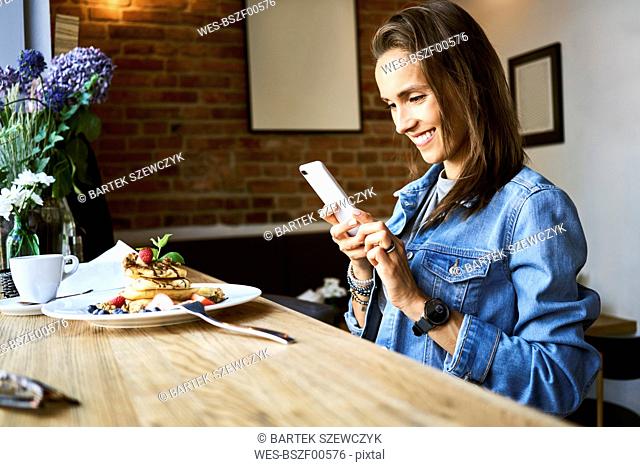Smiling young woman with plate of pancakes using phone in cafe