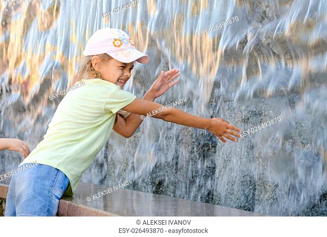 Cheerful six year old girl in summer clothes hand trying to get the water artificial waterfall