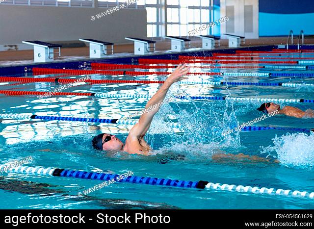 Side view of two Caucasian male swimmers at swimming pool, racing each other in lanes, swimming backstroke. Swimmers training hard for competition