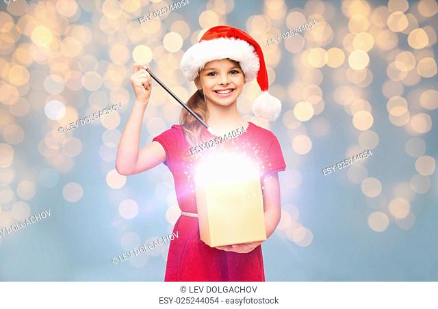 christmas, people, holidays and magic concept - smiling girl in santa helper hat with gift box and magic wand over lights background