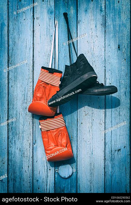 Black textile worn sneakers and red leather boxing gloves hanging on a blue wooden wall, vintage toning