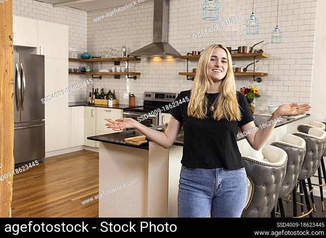 Portrait of a young woman standing in her home kitchen looking at camera with her arms out in a welcoming manner