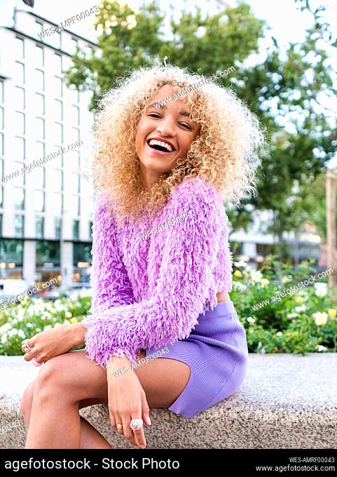 Happy young woman with blond Afro hairstyle sitting on bench