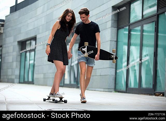 Young pair in love of stylish teenagers ride longboards tenderly holding hands on in the industrial background