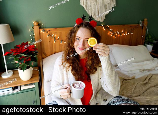 Smiling woman holding cup of hot chocolate with marshmallows sitting on bed at home