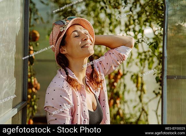 Smiling farmer wearing hat with eyes closed at doorway of greenhouse