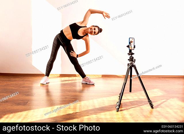 Online fitness trainer recording class of stretching, posing bending body with raised arm aside, wearing black sports top and tights
