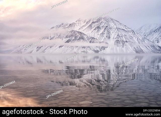 Snow covered Mount Worthington reflected in the still waters of Kathleen Lake with misty, low clouds and muted colors creating a moody atmosphere; Haines...