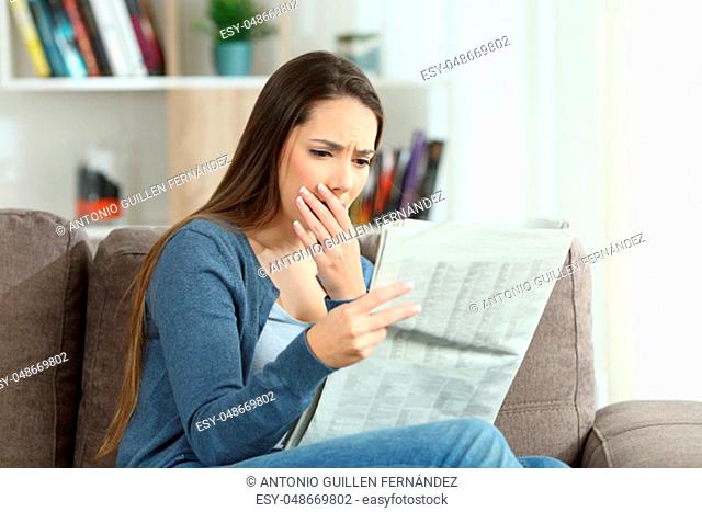 Worried woman reading bad news in a newspaper sitting on a couch in the living room at home