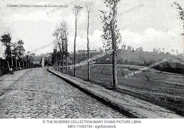 Cassel, France - Chemin de la Croix-Rouge (Red Cross Street). A cobblestoned street lined with trees and fields. There are several windmills visible on the...