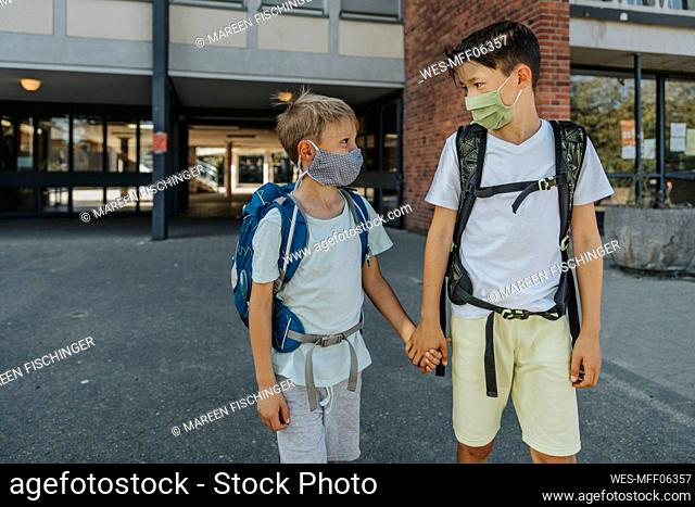 Brothers wearing protective face mask holding hands standing on street in front of school building