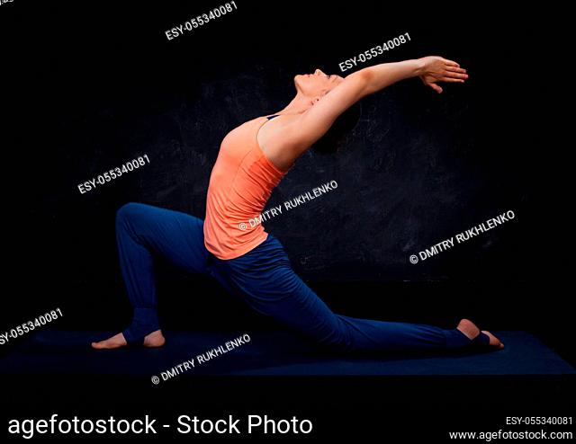 Beautiful sporty fit yogini woman practices yoga asana Anjaneyasana - low crescent lunge pose in surya namaskar on dark background