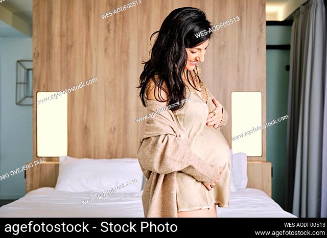Smiling pregnant woman with hands on stomach standing in bedroom at home
