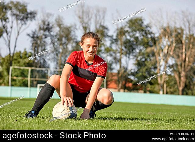 Happy girl with soccer ball crouching on field on sunny day