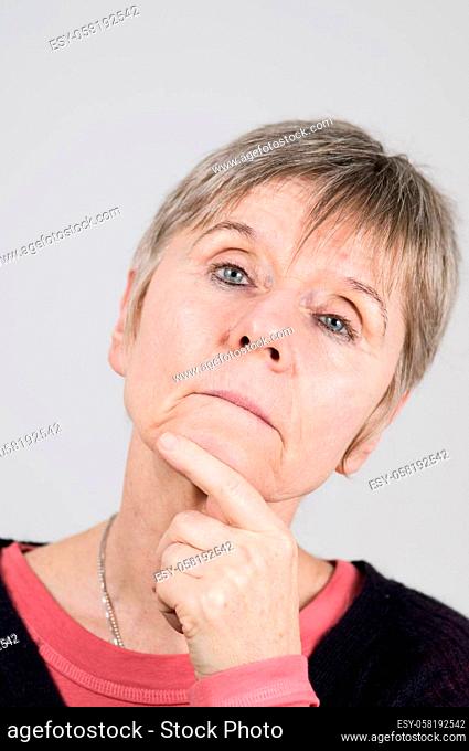 Head portrait of an older woman in front of a light gray background looking thoughtfully and skeptically at the camera with her left index finger on her chin