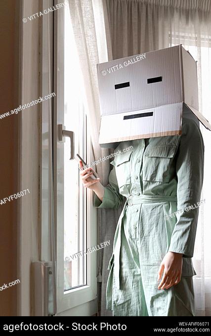 Woman wearing a cardbox on head with bored smiley looking out of the window and using smartphone