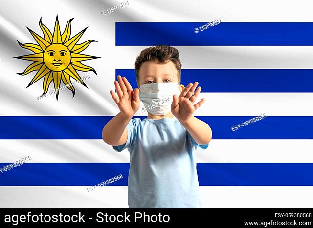 Little white boy in a protective mask on the background of the flag of Uruguay. Makes a stop sign with his hands, stay at home Uruguay