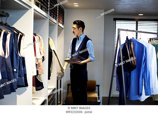 Japanese salesman with moustache wearing glasses standing in clothing store, looking at clothes on a rail, holding digital tablet