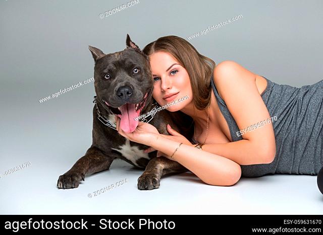 Beautiful sporty young woman lying on floor and hugging adult grey amstafford terrier dog. Studio shot over gray background. Copy space