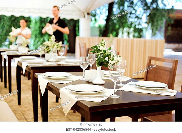 Summer terrace of the restaurant. On the tables laying with white plates of wine glasses and white vases with the colors of the bride