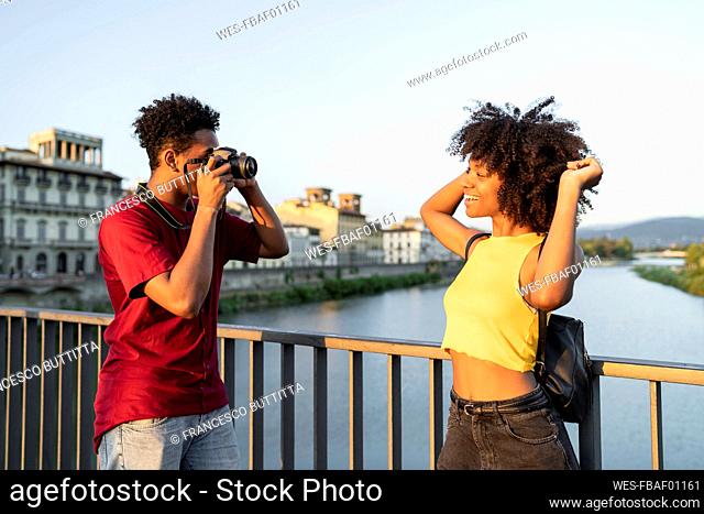 Young man taking a picture of his girlfriend on a bridge above river Arno at sunset, Florence, Italy