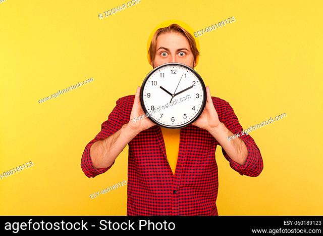 Portrait of amazed hipster guy in checkered shirt hiding face behind big clock and looking at camera with worried shocked expression, showing time to hurry up