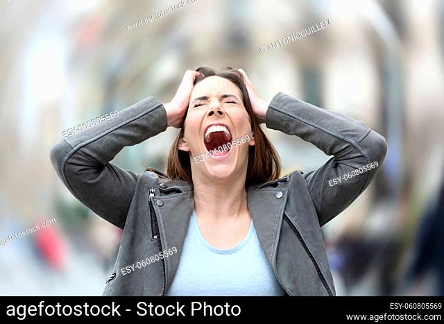 Front view portrait of a stressed woman screaming desperate suffering anxiety attack grabbing her head on city street