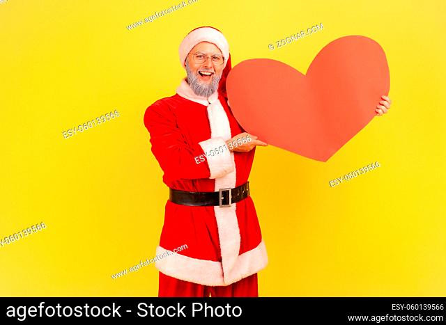Satisfied elderly man with gray beard wearing santa claus costume holding red heart in hands, expressing sensitive feelings, looking at camera