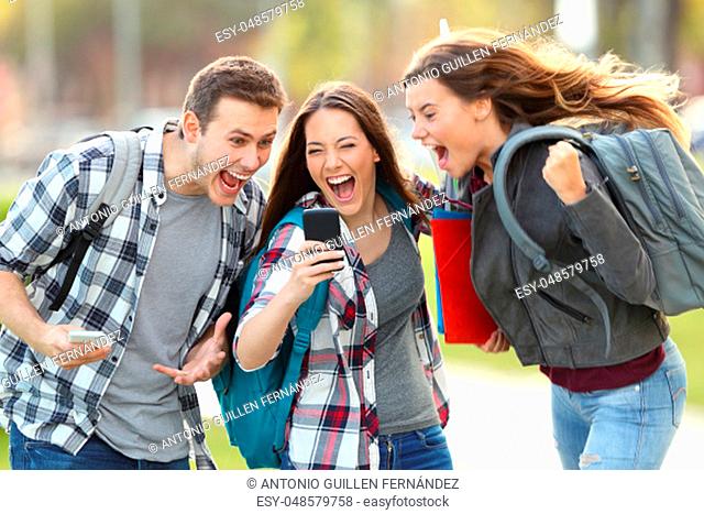 Front view of three excited students receiving good news on line in a mobile phone in an university campus or street