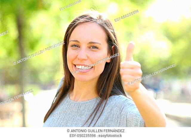 Happy woman looking at camera with thumbs up standing outdoors in a park