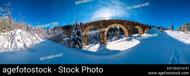 Stone viaduct (arch bridge) on railway through mountain snowy fir forest. Snow drifts on wayside and hoarfrost on trees and sunshine in sky