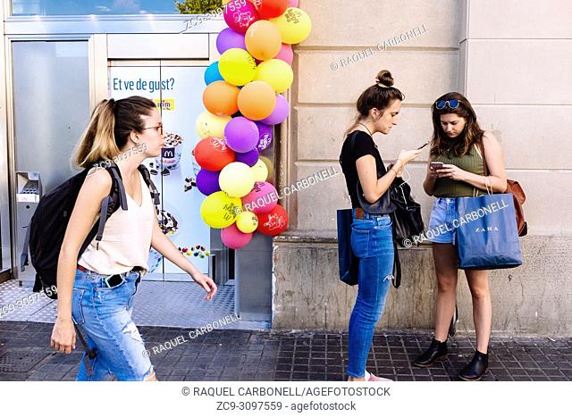 Young women looking at mobile phones next to colourful McDonald's ballons while other young woman carrying mobile in pocket is passing in front of them