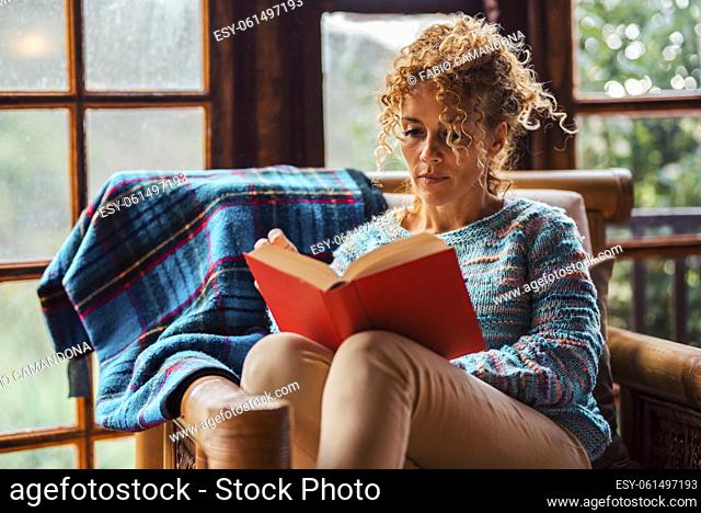 One serene woman at home relaxed sitting on a chair reading a book. Education and indoor leisure activity at home for happy single female people