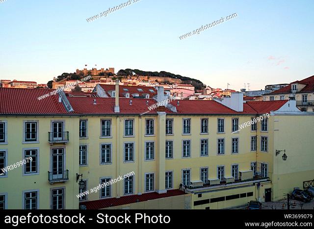 View of Lisbon Sao Jorge castle from Rossio train station with yellow building on the foreground at sunset, in Portugal