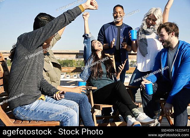 Group of young co-workers hanging out on rooftop patio having a drink and celebrating achievement reached on cell phone