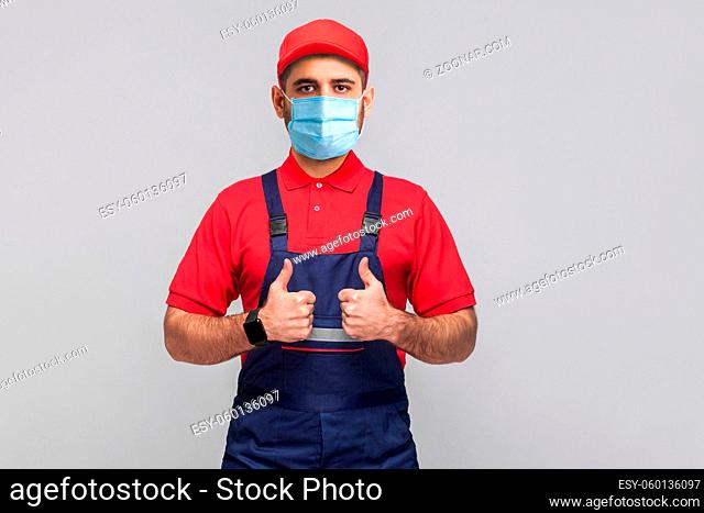 Work are done! Portrait of young man with surgical medical mask in blue overall, red t-shirt , cap, standing and showing thumps up and looking at camera
