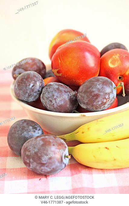 Plums and a small green apple in a white ceramic with bananas  Fresh purple plums in an white bowl  Small green apple windfall  Bananas lay beside the bowl on...