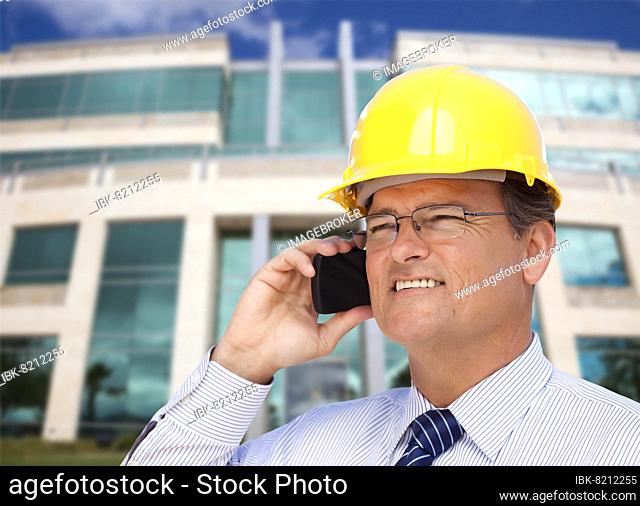 Handsome contractor in hardhat and necktie smiles as he talks on his cell phone in front of building