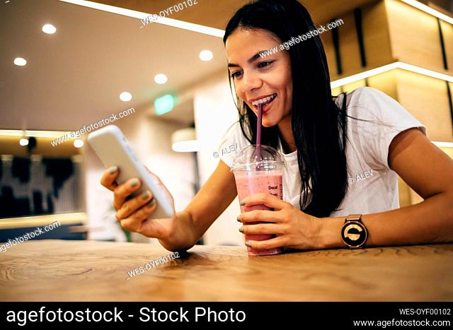 Black-haired woman drinking a smoothie and using smartphone in cafe