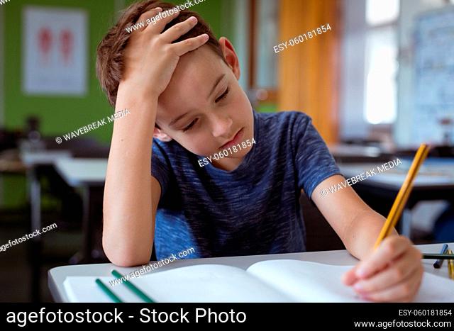 Tired caucasian schoolboy in classroom sitting at desk, holding head and writing