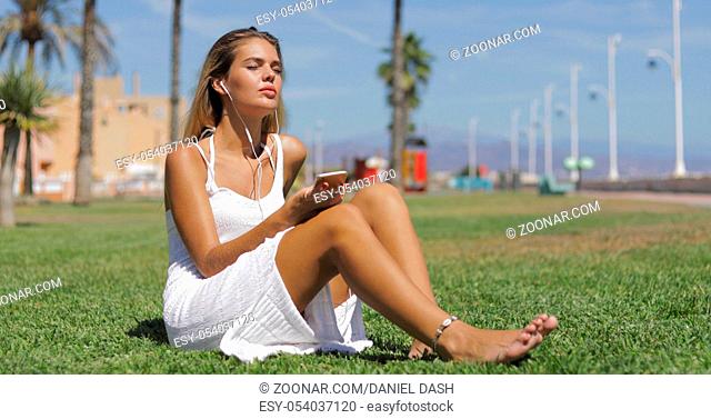 Young wonderful girl posing in white dress sitting barefoot on green meadow enjoying music with smartphone and headphones in bright tropical sunlight