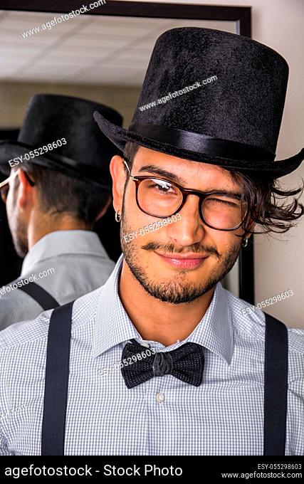 Portrait of brunette young man in glasses, hat, bow-tie, suspenders and shirt looking at camera. Mirror behind him