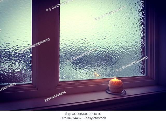 Candle burning near a frosted window in twilight. Winter evening
