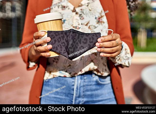 Woman holding bamboo mug and protective face mask standing in city on sunny day